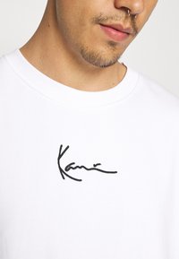 Witte katoenen T-shirt met een zwart geborduurd handtekeningontwerp. De ronde hals heeft een geribbelde textuur.