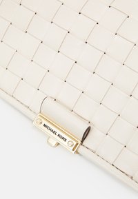 Gros plan sur le coin d’un sac à main en cuir tressé blanc avec un fermoir doré gravé « MICHAEL KORS » sur fond blanc.