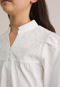 Bílá bavlněná halenka s jemnou výšivkou, mandlovým límečkem s volánkem a třemi knoflíky na légu. Měkká textura, volný střih.