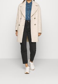 Kvinde iført en kridhvid struktureret frakke over en blå denimskjorte, sorte jeans og hvide sneakers, der står foran en ensfarvet baggrund.