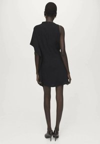 Zwarte jurk tot op de knie met asymmetrisch schouderontwerp, gestructureerde stof en ritssluiting aan de achterkant, gecombineerd met zwarte hakken.