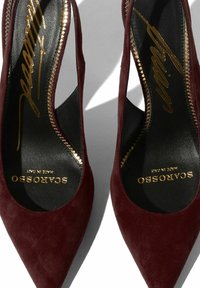 Scarpe con tacco alto in suede bordeaux, con punta affilata, lati intagliati e suole nere con lettere dorate. Dettagli di cuciture testurizzate visibili.