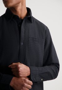 Zwart textuur overhemd met knopen, voorzien van een subtiel stippenpatroon, een borstzak en knoopmanchetten met een lichte glans.