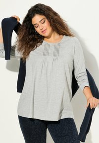 Hellgraues Langarmoberteil mit plissierten Akzenten, kombiniert mit einer marineblauen gerippten Strickjacke. Dunkel gemusterte Leggings runden das Outfit ab.