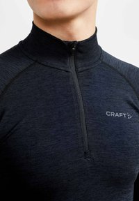 Zwarte, langärmelige top met halverwege ritssluiting, gemaakt van een gestructureerde stof. Heeft een opstaande kraag en een klein "CRAFT" logo op de borst.