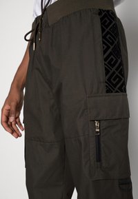 Cargo-Hosen in dunklem Olivgrün mit strukturiertem schwarzem Seitenpaneel, elastischem Bund und Reißverschlusstaschen-Details.