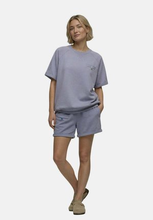 Femme debout, une main dans la poche, portant un haut à manches courtes violet clair et un short assorti avec des chaussons sur un fond uni.