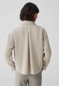 Light beige, long-sleeve shirt with vertical ribbed texture and shirt collar, featuring a back pleat and rounded hem.