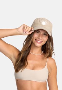 Beige corduroy pet met een witte zonafbeelding en tekst, scheef gedragen, met een gebogen rand en een verstelbare band aan de achterkant.