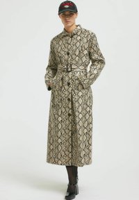 Bež in črna plašč s poskokom, z oblečenim pasom, gumbno zaporo in stranskimi žepi, nosi s črno kapo in robustnimi čevlji.