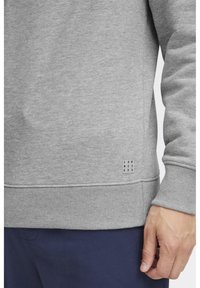 Grå hettegenser med ribbet mansjetter og nederkant. Har et svart logo-lapp nær bunnen. Stoffet ser mykt og teksturert ut.