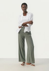 Pantalons olive à jambes larges associés à un débardeur blanc et une chemise en lin blanche déboutonnée. La tenue est complétée par des tongs simples.