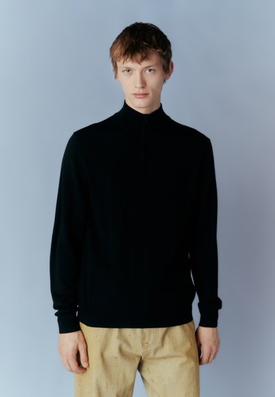 Μαύρο φούτερ με φερμουάρ και μακριά μανίκια, κατασκευασμένο από μαλακό υλικό. Διαθέτει ψηλό γιακά και στενή εφαρμογή. Συνδυάζεται με παντελόνι ανοιχτού χρώματος.