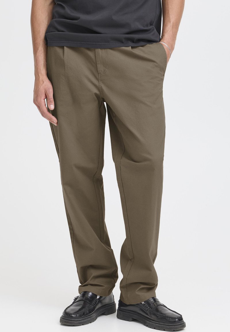 Pantalon vert olive à coupe droite, en tissu lisse. Comprend deux poches latérales et porté avec des chaussures noires. Design simple et fonctionnel.