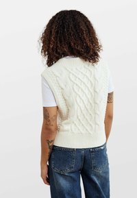 Cremfarbener, gestrickter ärmelloser Pullover mit Kabelmustern und geripptem Saum, getragen über einem weißen Hemd, kombiniert mit blauen Jeans.