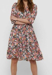 Mujer vestida con un vestido floral hasta la rodilla en tonos rosa, naranja y verde, de pie con los brazos cruzados frente a un fondo liso.