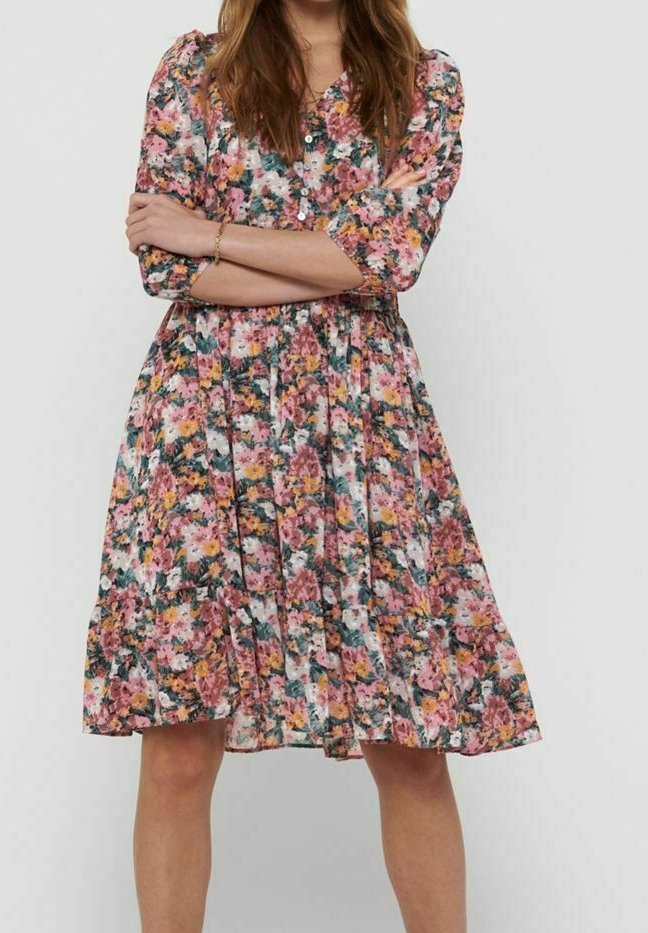 Mujer vestida con un vestido floral hasta la rodilla en tonos rosa, naranja y verde, de pie con los brazos cruzados frente a un fondo liso.