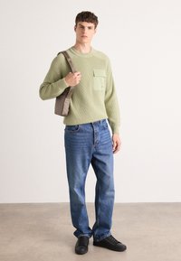 Un pull côtelé vert clair avec une poche poitrine, associé à un jean en denim bleu et des baskets noires. La silhouette tient un sac à bandoulière marron.