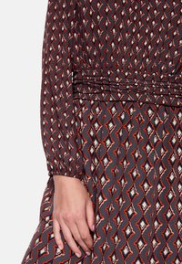 Langärmeliges Kleid aus dunklem Stoff mit einem geometrischen Muster aus roten, weißen und blauen Rauten. Verfügt über einen gerafften Taillendetail und Falten.