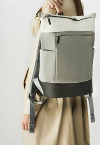 Zaino grigio e nero con parte superiore crema, dotato di una tasca con zip, lato in rete e spallacci regolabili. Accenti in tessuto a trama visibili.