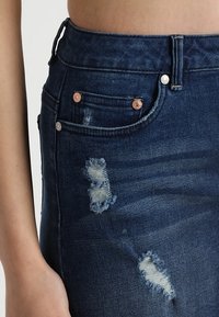 Tmavomodré džíny z denimu s vysokým pasem, s oprýskanými detaily a měděným kováním na kapsách a pase.