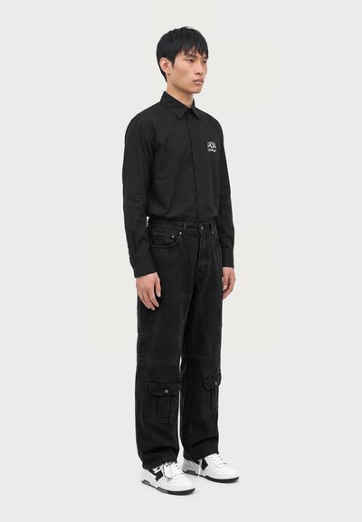 Jeune homme debout portant une chemise noire à manches longues boutonnée, un pantalon cargo noir avec poches et des baskets blanches sur un fond uni.