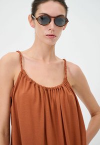 Top marrone senza maniche con scollatura arricciata e spalline intrecciate. Modello che indossa occhiali da sole rotondi con lenti scure, su uno sfondo semplice.