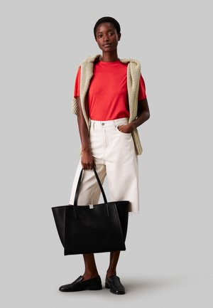 Mujer con camiseta roja, pantalones cortos blancos hasta la rodilla, suéter de punto beige sobre los hombros, mocasines negros, sosteniendo un bolso tote grande negro.