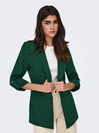 Žena s dugom tamnom kosom, odjevena u tamnozeleni sako, bijelu majicu i bež hlače, drži rever saka.