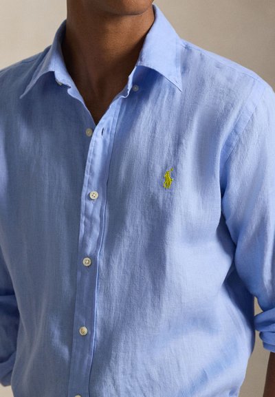 Homme portant une chemise bleu clair boutonnée avec un petit logo jaune brodé sur la poitrine gauche, manches retroussées.