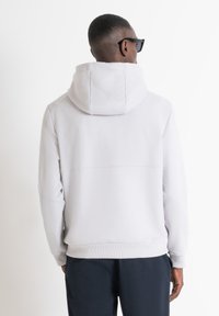 Sudadera con capucha de color gris claro, con un bolsillo canguro, puños y dobladillo acanalados; textura suave. Modelo visto desde la parte de atrás, usando gafas de sol.