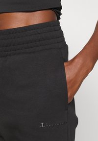 Pantalones de chándal negros hechos de un tejido suave, con una cintura de canalé, bolsillos laterales y el logotipo de "Champion" bordado en la parte delantera.