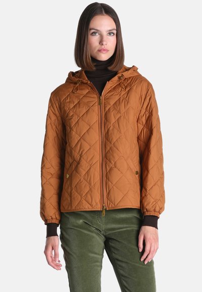Femme portant une veste matelassée marron zippée avec capuche par-dessus un col roulé noir et un pantalon vert, debout devant un fond clair uni.