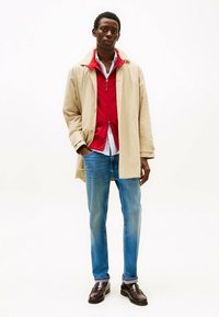 Cappotto leggero beige, giacca rossa con zip sotto, camicia bianca, jeans in denim blu e mocassini marrone scuro; il modello si trova davanti a uno sfondo bianco.