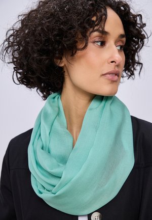 Frau mit lockigem dunklem Haar, die einen helltürkisen Schal und ein schwarzes Oberteil trägt, schaut nach rechts vor einem schlichten, hellen Hintergrund.