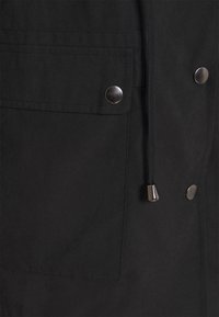 Svart jacka med en framficka som har en silverknapp och en dragsko med metallspetsar. Strukturerad tyg.