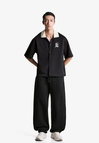 Zwarte button-up shirt met een crèmekleurige kraag en een prominent "K"-logo. Gecombineerd met losse zwarte broeken en witte sportschoenen.