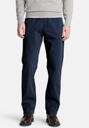 Hombre con pantalones azul marino de pierna recta, suéter gris y zapatos marrón oscuro con cordones de pie sobre un fondo blanco.