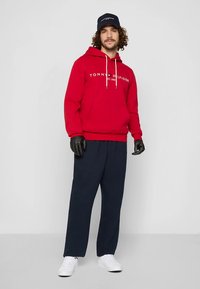 Rode capuchontrui met witte "TOMMY HILFIGER"-logo, gecombineerd met donkerblauwe broek met losse pasvorm en witte sneakers. Handschoenen en pet inbegrepen.