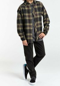 Camicia di flanella a quadri nera, verde e gialla; t-shirt grigia sotto. Pantaloni neri e scarpe nere con dettagli turchesi.