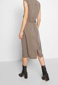 Knielanges, taupefarbenes Kleid mit tailliertem Bund, kurzen Ärmeln und Schlitzdetails im Rücken, kombiniert mit schwarzen und roten Knöchelboots. Strukturierter Stoff.