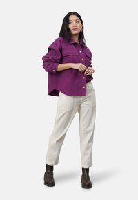Fioletowa koszula zapinana na guziki z dużymi kieszeniami i podwiniętymi rękawami, zestawiona z kremowymi spodniami z kordurowej tkaniny i czarnymi botkami do kostki. Prosty, luźny fason.