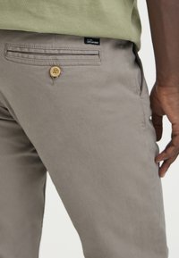 Pantaloni di cotone grigio chiaro con tasca posteriore, chiusura con bottone e etichetta visibile. Texture liscia e vestibilità sartoriale.
