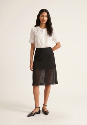 Kvinde med langt mørkt hår iført en hvid kortærmet bluse og sort gennemsigtig midi nederdel med blondekant, stående mod en ensfarvet baggrund.