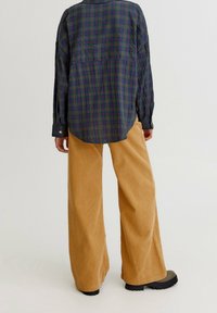 Chemise en flanelle à carreaux bleu marine et vert à manches longues, associée à un pantalon en velours côtelé à jambes larges de couleur fauve, présentant une texture lisse.