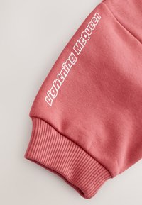 Gros plan d'une manche de sweat-shirt rose avec poignet côtelé et texte blanc "Lightning McQueen" le long du bras.