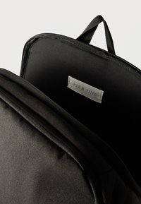 Εσωτερικό μαύρης υφασμάτινης τσάντας πλάτης με λεία υφή, κλείσιμο με φερμουάρ και ετικέτα που γράφει «PIER ONE» σε ανοιχτό μπλε πάνω σε λευκό φόντο.