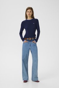 Woman standing with hands in pockets wearing navy ribbed sweater, wide-leg light blue jeans, brown belt, and burgundy pointed shoes.