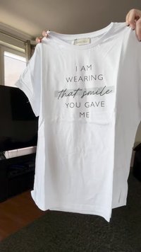 T-shirt branco segurado por mãos com o texto "Estou a usar esse sorriso que me deste" em letras pretas.
