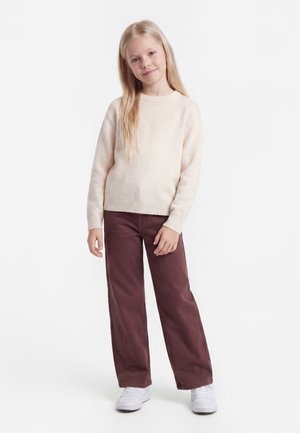 Ragazza giovane in piedi con la mano sulla coscia, indossa un maglione beige, pantaloni marroni a gamba larga e sneakers bianche su uno sfondo bianco semplice.